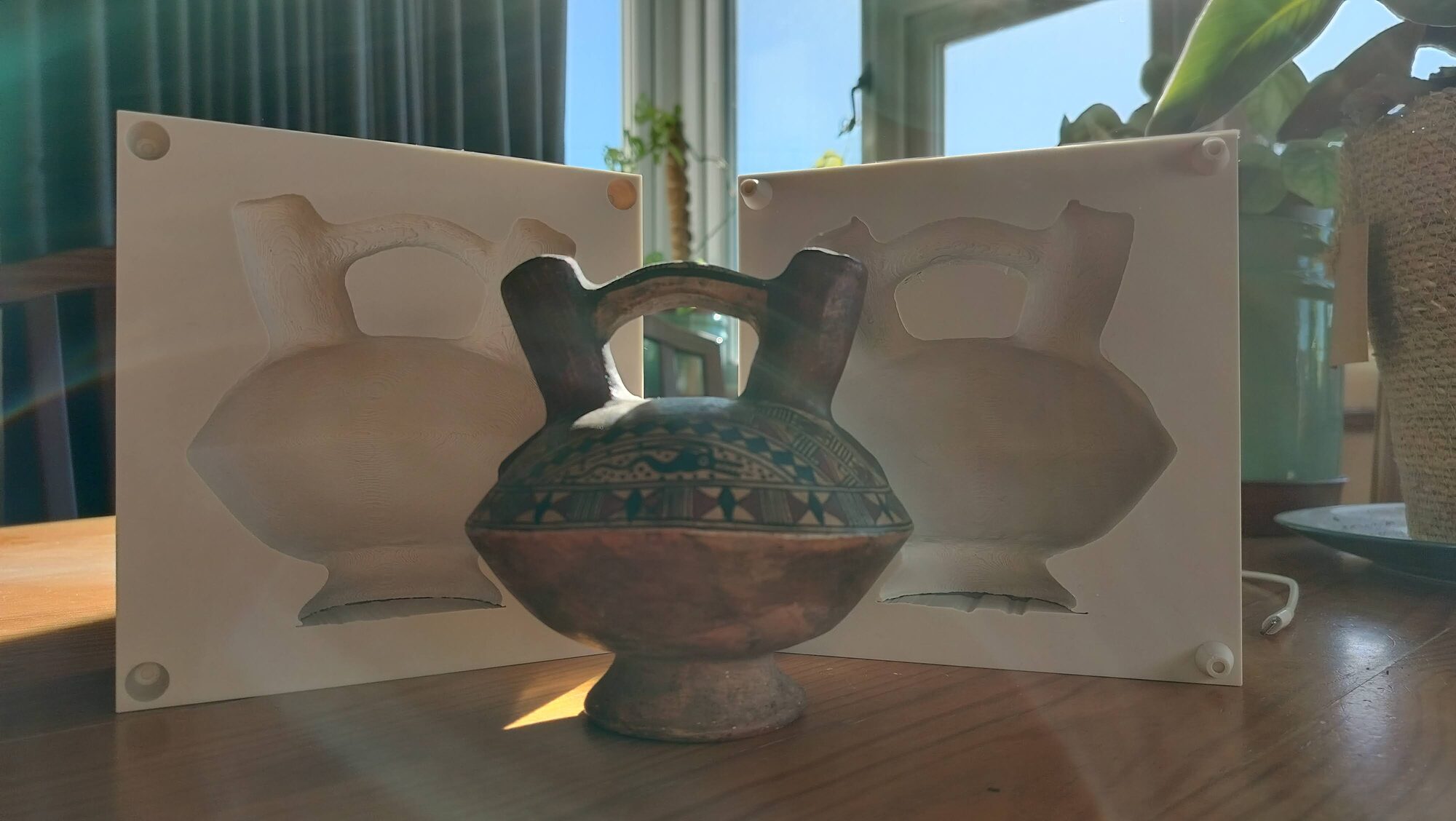 A painted double-spout-and-bridge ceramic vessel standing between the two open halves of its 3D-printed case, each half holding a cavity that matches the vessel's outline